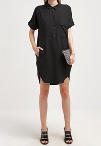 Abito camicia nero a maniche corte con colletto, chiusura a bottoni e una tasca sul petto. Tessuto testurizzato con spacco laterale. Accessorio clutch nero.