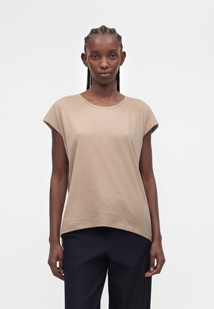 Femme aux cheveux tressés portant un haut beige à manches courtes et un pantalon noir, debout sur un fond blanc uni.
