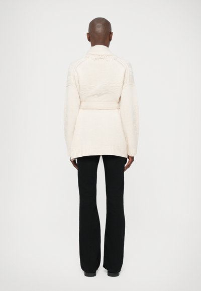 Cardigan creme de mangas longas com um cinto na cintura, padrão texturizado nos ombros e um fundo ligeiramente alongado, combinado com calças pretas.