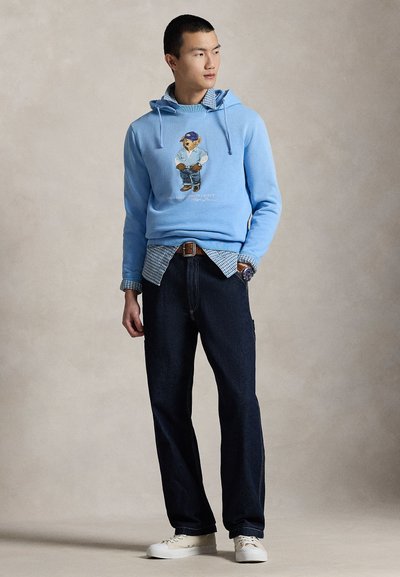 Mladić u svijetloplavoj dukserici s grafikom medvjeda, preko koje nosi kariranu košulju, tamne traperice i bijele tenisice, stoji i gleda lijevo.