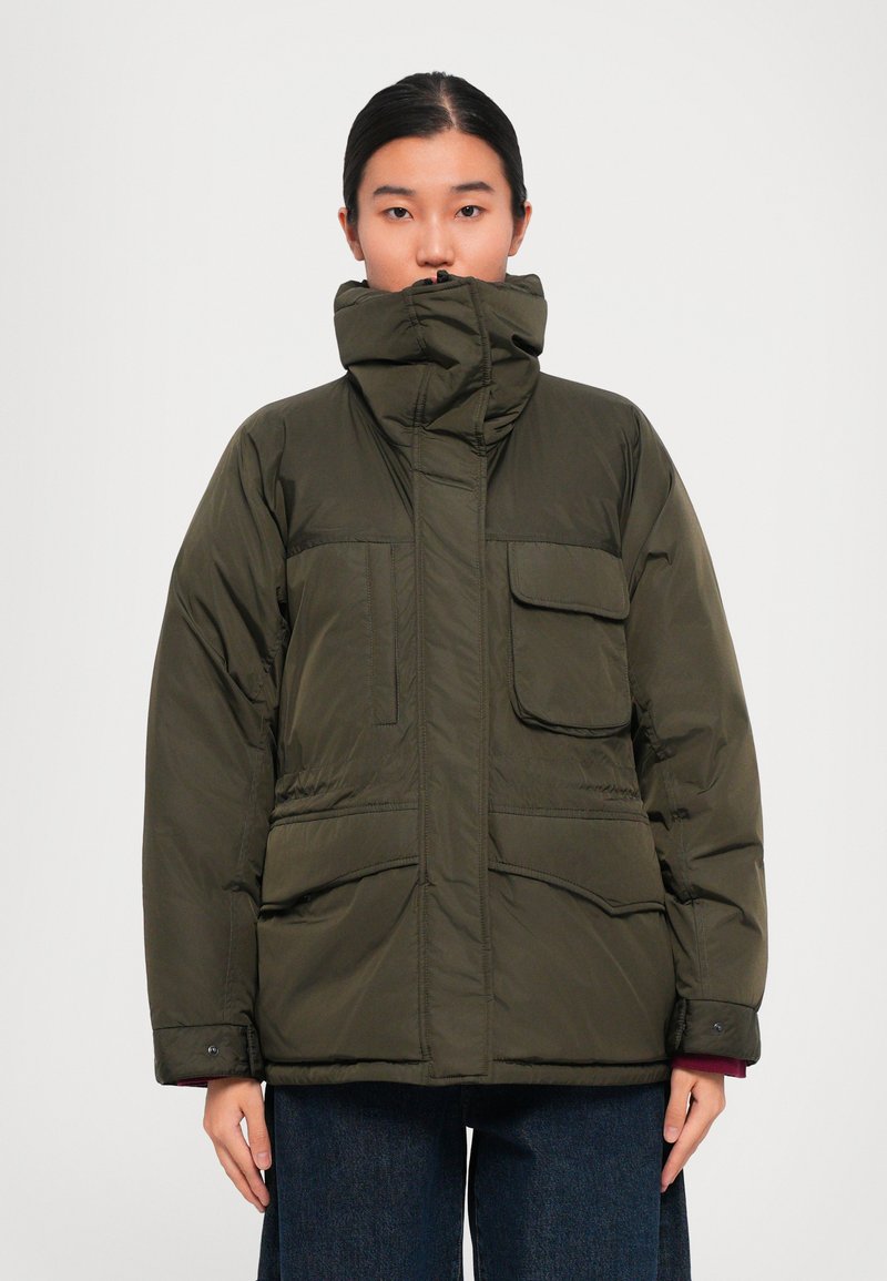 Person wearing an olive green padded jacket with high collar and multiple large front pockets, paired with dark blue pants.