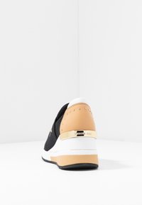 Basket noire avec une tige en mesh texturé, accent beige au talon, détail en métal doré et une semelle intermédiaire blanche. Fond de studio.