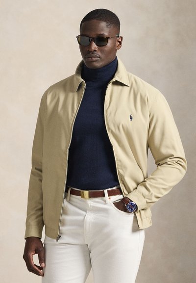 Mand iført beige jakke, navy højhalset trøje, hvide bukser, brun bælte, blå ur og mørke solbriller på ensfarvet baggrund.