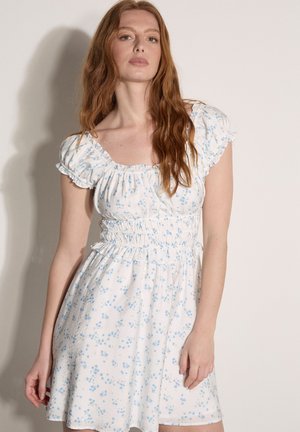 Kobieta z długimi rudymi włosami w białej sukience z niebieskim kwiatowym wzorem i bufkami na krótkich rękawach na jednolitym tle.