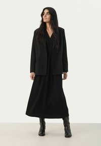 Schwarzer Blazer über einem schwarzen gemusterten Maxikleid. Das Kleid hat ein strukturiertes Design und das Outfit wird mit schwarzen kniehohen Stiefeln kombiniert.