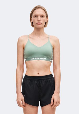Femme aux cheveux blonds courts portant un soutien-gorge de sport Jordan vert clair et un short de sport noir Nike, debout face à l'avant sur un fond uni.