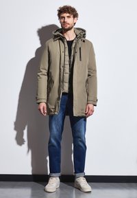 En khaki parka med huva, dragkedja och tryckknappar, sidofickor, lager över en svart topp, matchad med blå jeans och beige sneakers.