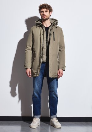 Khaki parka med hætte, lynlås og knaplukninger, sidelommer, lagdelt over en sort top, parret med blå jeans og beige sneakers.