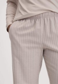 Person in beigen gestreiften Hosen mit elastischem Bund und einem beigen Langarmshirt, entspannt die Hand in der Tasche.