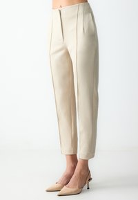 Beige op maat gemaakte broeken met een hoge taille, voorzien van twee voorplooien en een taps toelopend been. Gemaakt van een soepele, gestructureerde stof, met zijzakken.