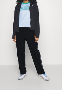 Sort atletisk jakke, lyseblå mesh t-shirt med "NIKE" tekst, sorte bukser med sidelister og grå sneakers med blå accenter.