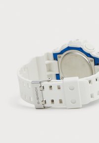 Montre G-SHOCK avec un bracelet en caoutchouc blanc présentant des motifs découpés. Accents métalliques bleus autour du boîtier et un fond en acier inoxydable.