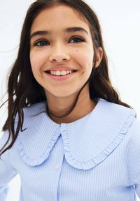 Sininen ja valkoinen raidallinen paita, jossa on rypytetty, ylisuuri kaulus. Napitus edessä, pehmeä kangas ja rento muotoilu.