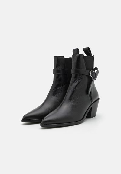 Bottines noires en cuir avec un bout pointu, un talon moyen, et un détail de sangle avec une boucle en argent sur le côté. Texture lisse.