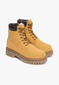 Gelbe Wildleder-Schnürstiefel mit einem braunen gepolsterten Kragen, dickem Sohlenmaterial und metallischen Ösen für die Schnürsenkel. Texturierte Oberfläche, robuste Bauweise.