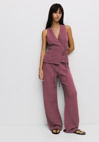 Tvådelad outfit i texturerad, mauve-färgad tyg. Toppen har en ärmlös omslagdesign med sidofäste, ihopparad med vida byxor. Platt sandaler.