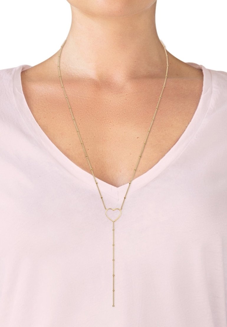 Collier chaîne en or avec un pendentif en forme de cœur et une chaîne pendante portée sur un t-shirt rose clair à col en V sur la poitrine d'une personne.