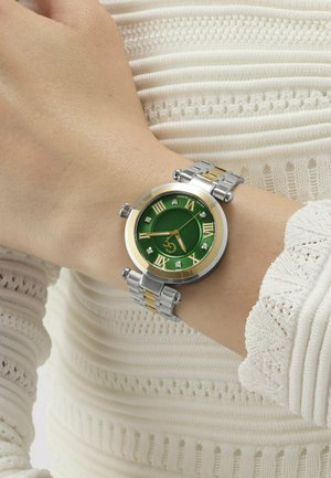 Montre-bracelet avec cadran vert, chiffres romains dorés, et bracelet en métal argenté-doré portée sur le poignet gauche par-dessus un haut à manches longues blanc texturé.