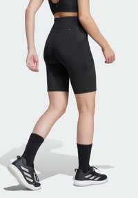 Shorts de sport noirs avec une taille haute, fabriqués en tissu lisse. Présentent un design épuré et des accents sur les panneaux latéraux. Portés avec des chaussettes et des chaussures noires.