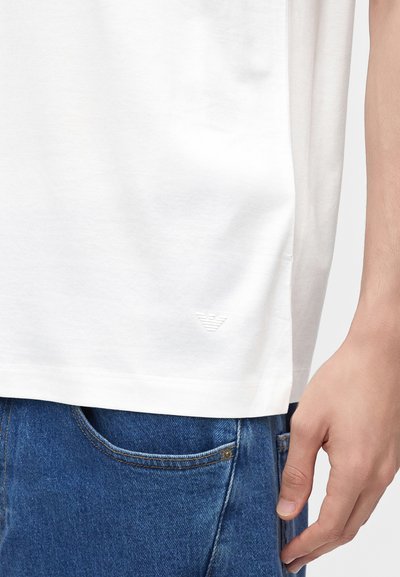 Κοντινό πλάνο ενός ατόμου που φοράει λευκό T-shirt και μπλε τζιν, με ένα μικρό ανάγλυφο λογότυπο κοντά στο τελείωμα του T-shirt και ένα χέρι στο πλάι.