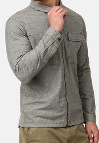 Homme portant une chemise gris clair à fermeture éclair avec poches poitrine à rabat et poignets boutonnés, ajustant le col d'une main.