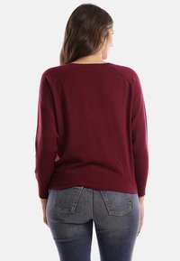 Donna con capelli castano lunghi che indossa un maglione bordeaux a maniche lunghe e jeans blu, vista di spalle su sfondo bianco.