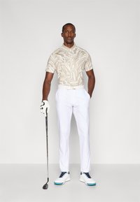 Beiges Poloshirt mit abstrakten weißen Mustern, kombiniert mit weißen Hosen. Die Accessoires beinhalten einen Handschuh und einen Golfschläger; das Schuhwerk hat blaue Akzente.