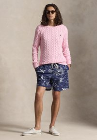 Jongere met ronde zonnebril, roze kabelgebreide trui, marineblauwe zwembroek met bloemenmotief en witte sneakers, staand met handen in de zakken.