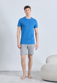 Άνδρας όρθιος με γυμνά πόδια σε παρκέ δαπέδου, φοράει μπλε μπλουζάκι και γκρι σορτς δίπλα σε καμπύλη καρέκλα με μπεζ ταπετσαρία.