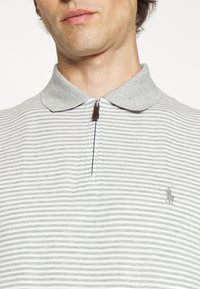 Grårandig pikétröja med grå krage, kort dragkedja och en liten broderad emblem på vänster bröst. Texturerat tyg.