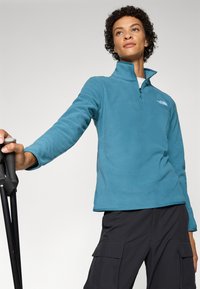 Teal fleece quarter-zip top with a stand collar, featuring a subtle logo on the chest. Model wears dark cargo pants and holds trekking poles.