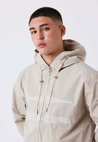 Giovane uomo con capelli corti che indossa una giacca beige con cappuccio, con una stella bianca e le lettere PXLP ricamate sul davanti.