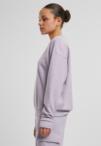 Felpa viola chiara con vestibilità morbida, polsini e orlo a costine, e scollo rotondo, indossata da una persona di profilo.