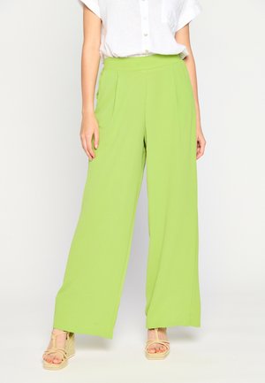 Mujer que lleva pantalones anchos de color verde brillante combinados con una camisa blanca de manga corta con botones y sandalias beige de cuña.