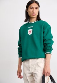 Grønn hettegenser med rund hals, kontrastfargede hvite striper og et brodert logo-patch på brystet. Kombinert med stripete bukser.