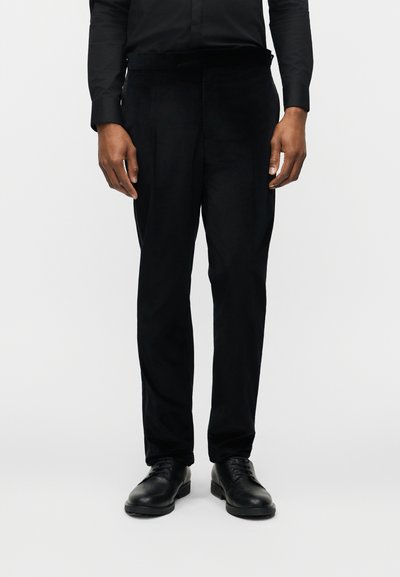 Άνδρας που φοράει μαύρο επίσημο παντελόνι, μαύρα επίσημα παπούτσια και μαύρο πουκάμισο με μακριά μανίκια, στέκεται μπροστά σε λευκό απλό φόντο.