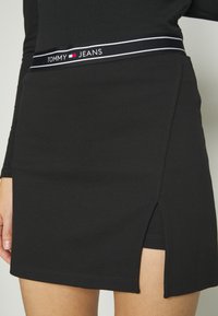 Žena oblečená v černé sukni se slabým rozparkem a elastickým páskem značky Tommy Jeans přes odpovídající černé šortky.