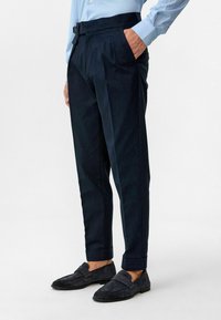 Marineblauwe chino broeken met een getailleerde pasvorm, voorzien van een riem en zijzakken. De stof is gestructureerd, met een gladde afwerking en opgerolde manchetten.