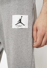 Grå sweatpants med en mjuk textur, med en vit patch som visar en svart "Jumpman"-logotyp och "Flight"-text. Nära bild av fickområdet.