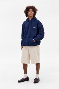 Felpa blu navy con un dettaglio testuale bianco, abbinata a pantaloni corti crema a righe sottili e mocassini scuri con calzini bianchi. Sfondo bianco semplice.
