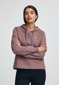 Sudadera rosa hecha de material suave, con un bolsillo frontal y capucha con cordón. Combinada con pantalones negros, el modelo muestra un ajuste relajado.