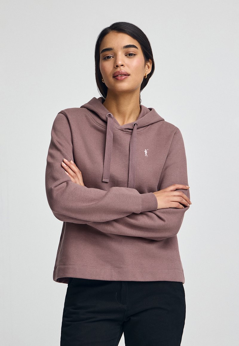 Sudadera rosa hecha de material suave, con un bolsillo frontal y capucha con cordón. Combinada con pantalones negros, el modelo muestra un ajuste relajado.