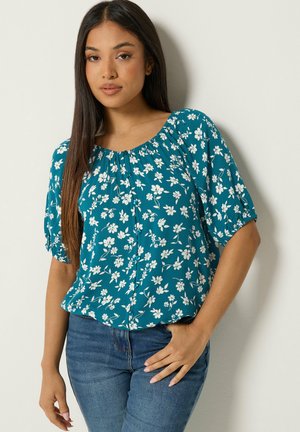 Mujer con el cabello largo y oscuro que lleva una blusa floral azul verdosa con mangas abullonadas y vaqueros azules, de pie frente a un fondo claro.