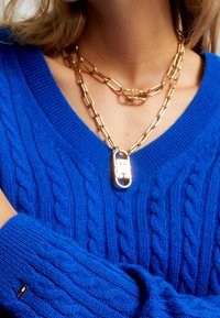 Pull bleu tricoté avec un motif torsadé, colliers en chaîne dorés superposés avec un pendentif cadenas, et un détail de marque sur la manche.