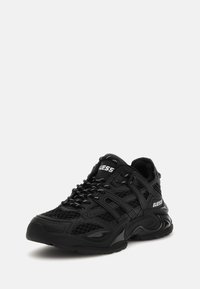 Baskets de sport noires avec une tige en mesh et en cuir, dotées d'une semelle en caoutchouc épaisse, d'accents texturés et d'un détail de logo blanc sur le côté.