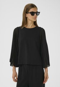 Top nero con maniche plissettate trasparenti, scollatura rotonda e vestibilità ampia. Il modello indossa occhiali da sole neri angolari per un contrasto aggiuntivo.