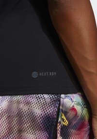 Close-up van de zijkant van een persoon met een donkere huid, die een zwart T-shirt draagt met het label "HEAT.RDY" en meerkleurige mesh shorts met zwarte stippen.