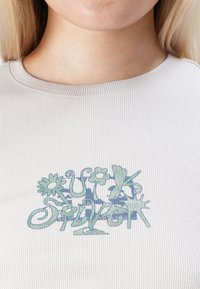 Suurendatud vaade ribatelje valgele särgile, millel on sinakasroheline lilleline kirjadi, mis ütleb "Just Super" naise lõua all.