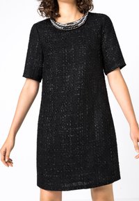 Robe noire à manches courtes en tissu texturé avec un col rond orné d'un collier en perles. Silhouette évasée, conçue jusqu'au genou.