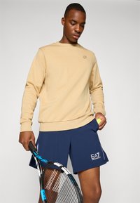 Beige genser med ribbestrikkede mansjetter og kant, parret med marineblå shorts med logo-design. Spiller som holder en tennisracket og ball.